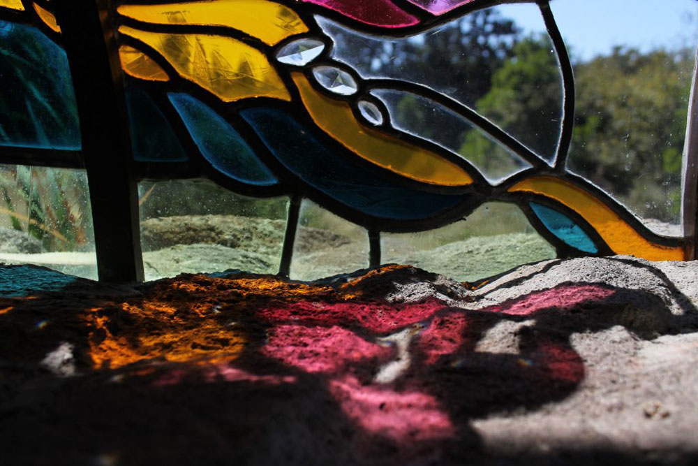 This image is a version of a self-portrait. In it is a stained glass window which is reflecting color onto the windowsill below it. Yellow, red, and blue light is laid across the bottom.