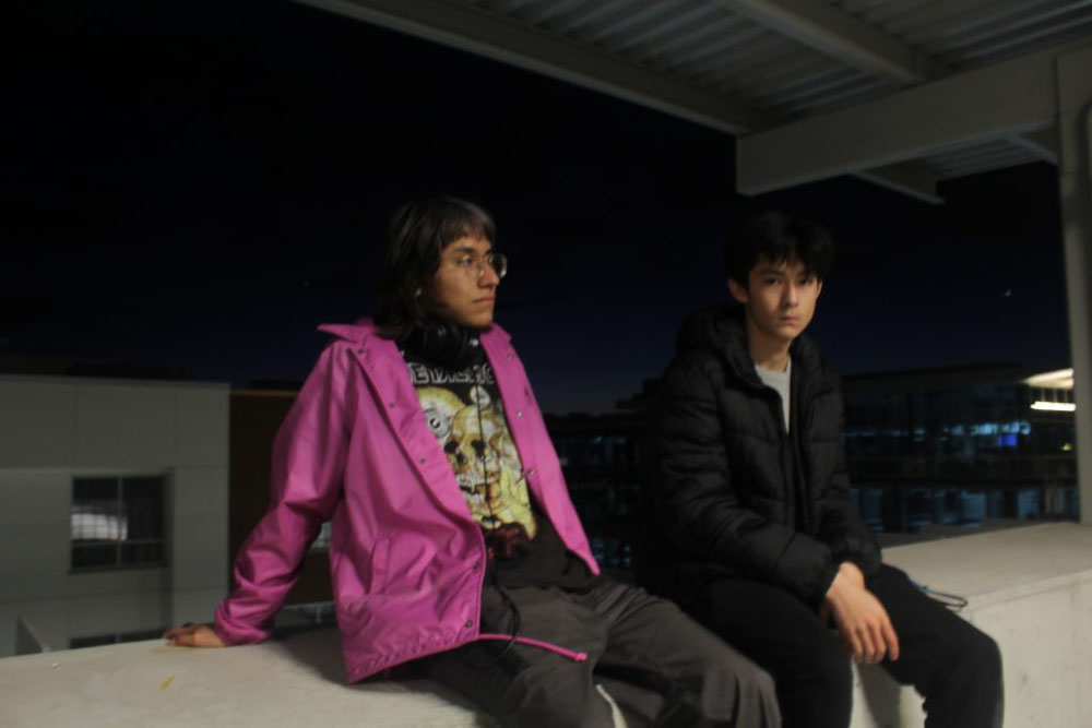 The photo is outdoors at the top level of a parking garage and background is dark. The main subjects are two male teenagers. One is looking at the camera and one is looking away. 