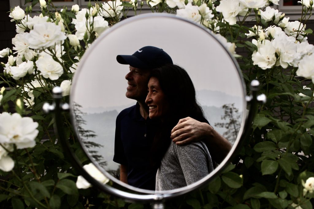 medium sized makeup mirror in front of a white rose bush. the mirror reflects a man and a woman smiling into the distance with a foggy sky and mountains in the background.