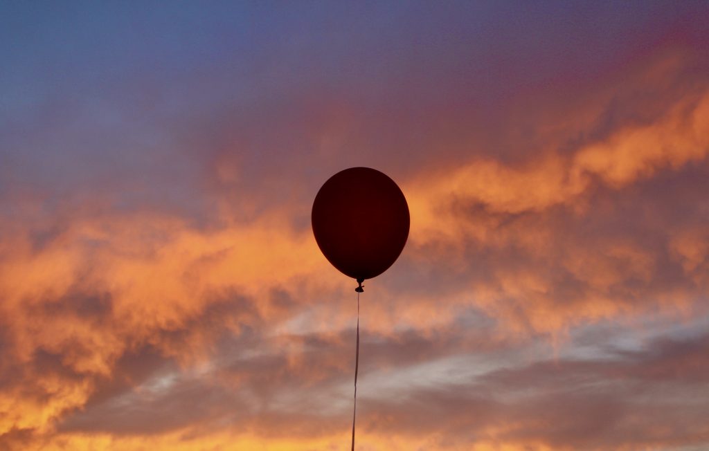 a red balloon backed by a vibrant sunset of blue, orange, yellow, and pink