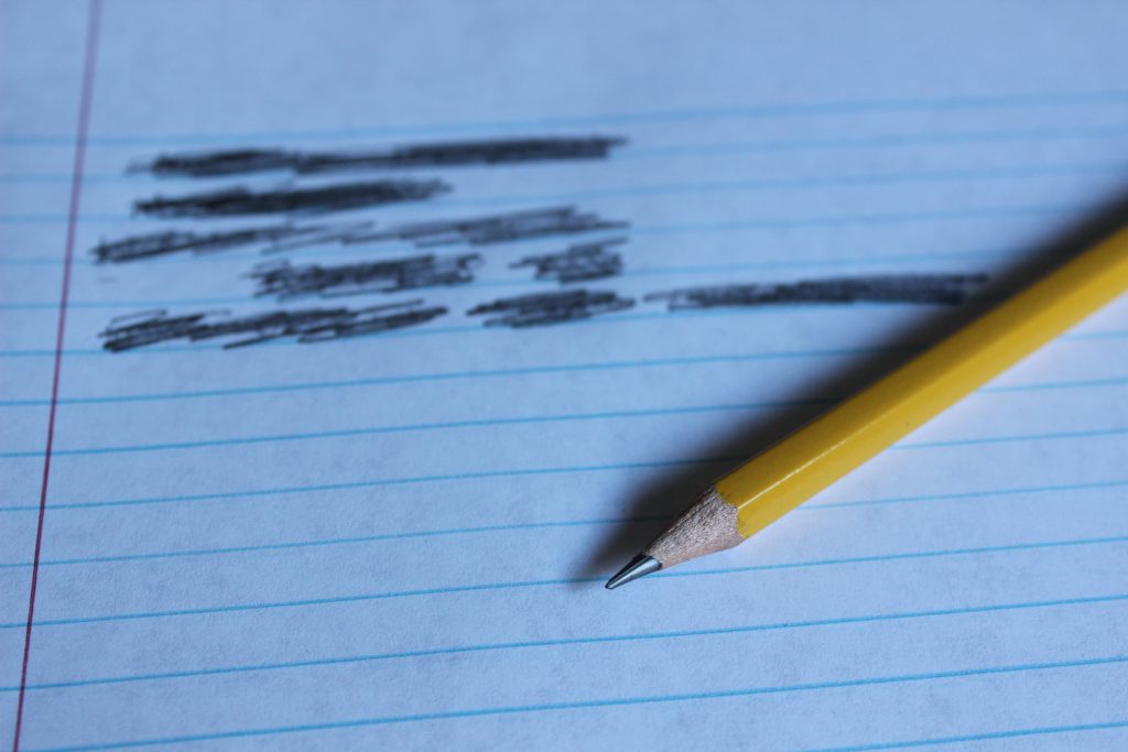 A pencil laying on a piece of line paper. There are a few lines that have crossed out writing on them.