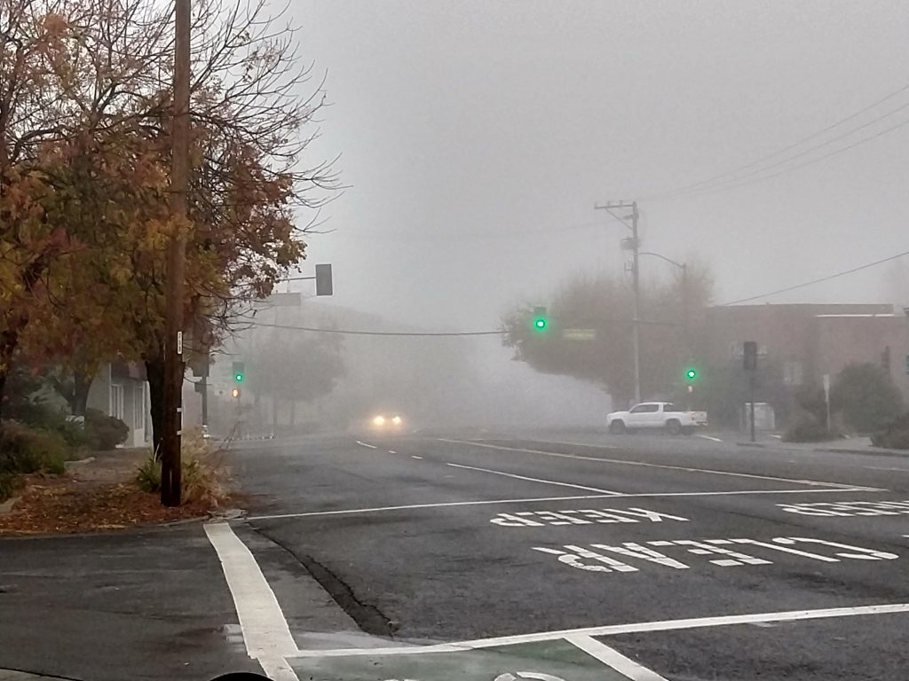 This picture shows a view of my hometown.  It is a foggy day, so distance visibility fades off.  The picture is of an intersection.  The lights facing the camera are green, and shine strangely through the fog.  There is a car approaching the perspective of the camera, though due to the fog, only the headlights are visible.