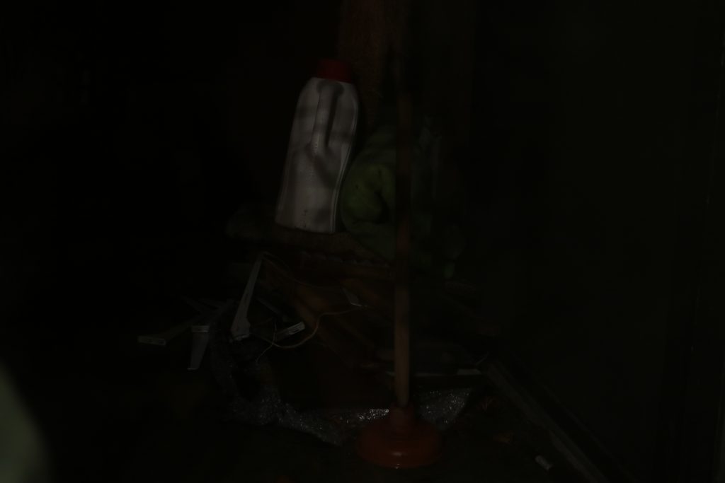 This is a picture of a pile of random trash, very darkly lit.  Visible on the pile is a white container of some sort, a plunger, and a rug.