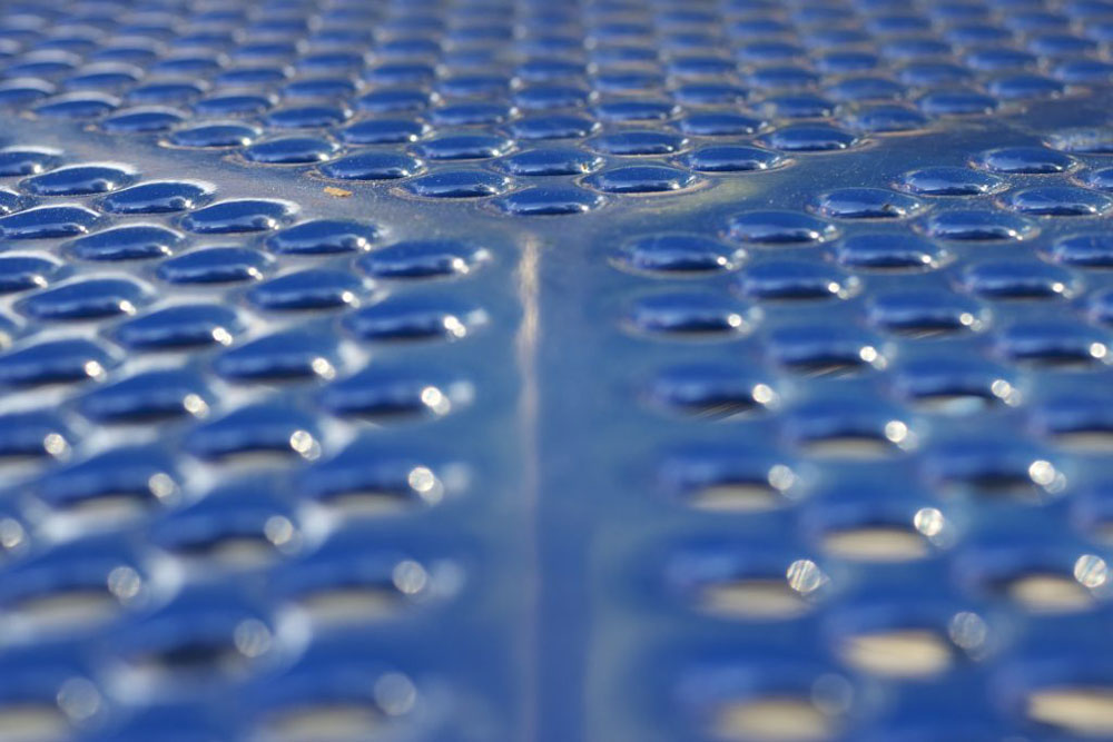A blue surface with many holes punched in it at even distances from each other is visible. However, there is a wide line of the blue surface, with no holes, leading form the bottom middle to the center of the photo. From there, two equally sized "lines" without holes diverge at 45 degree angle towards the upper left and right corners of the photo respectively.