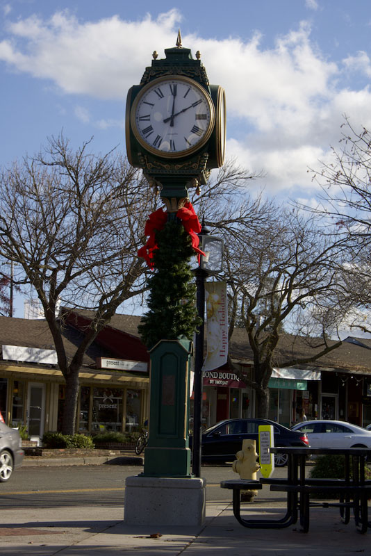 An image of a small clock tower in a town square. The clock is framed in gold on a green post, and there is a small wreath with a red ribbon on it. 