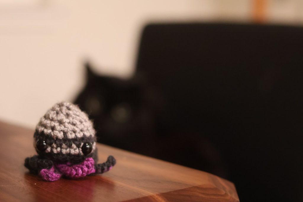 A handmade grey and purple octopus sitting on the corner of a desk. In the background, a blurry cat staring right at them on a chair.