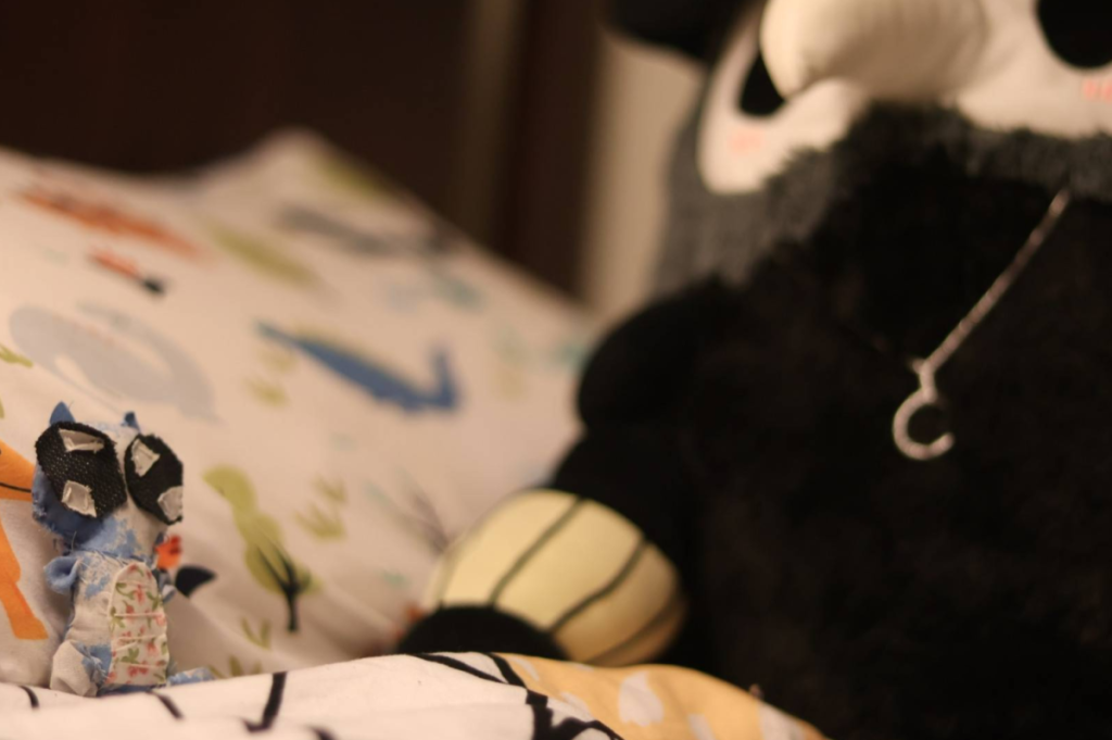 A small plushie baby dragon on the far left, sitting against a pillow and on a blanket. On the right, a giant black plushie in the shape of a plague doctor takes up half the image.