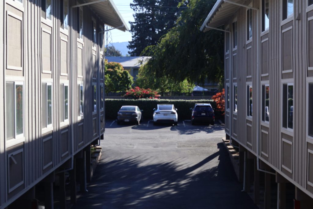An apartment parking lot, symmetrical buildings on the left and right side. In the distance, there is one white car in the middle and two black/dark ones on the right and left.