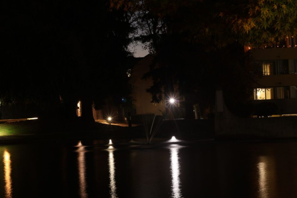A pond at night with a water fountain in the middle, the lights from the nearby lamps are reflected off the water. There are buildings and trees in the background.