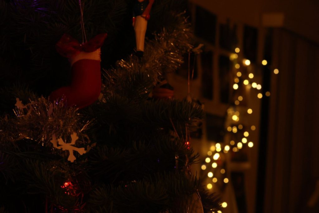 A christmas tree on the left with a sock shaped ornament as the main focus. In the background, there are blurred out yellow lights. Overall, the photo has an orange hue.