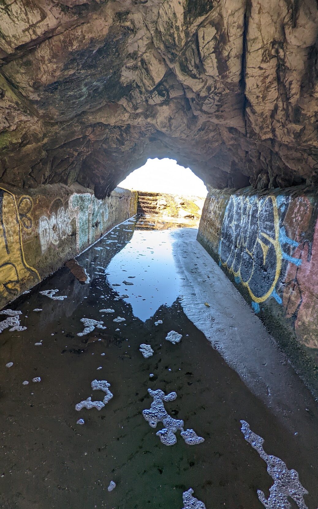 A picture of a tunnel with light on the other side. Graffiti lines the walls of the tunnel and there is water on the ground.