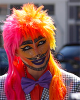 A drag king wearing a brightly colored wig, a holographic suit and a bowtie, and bold makeup that includes eyeliner and painted on facial hair.
