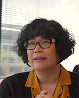 A photo of a woman with short curly dark hair, glasses, and a black sweater with a yellow shirt underneath.