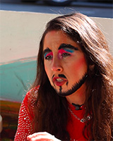 A photo of a drag king with long brown hair. They're wearing colorful makeup and a sparkly red outfit.