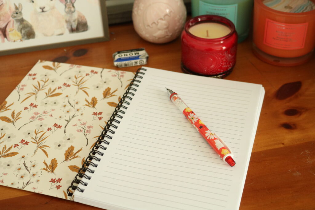 A photo of the first page of a notebook. There is nothing written on it, but a pencil sits, waiting to be used. (In the corner there are candles because the photographer thought it was aesthetic.) 