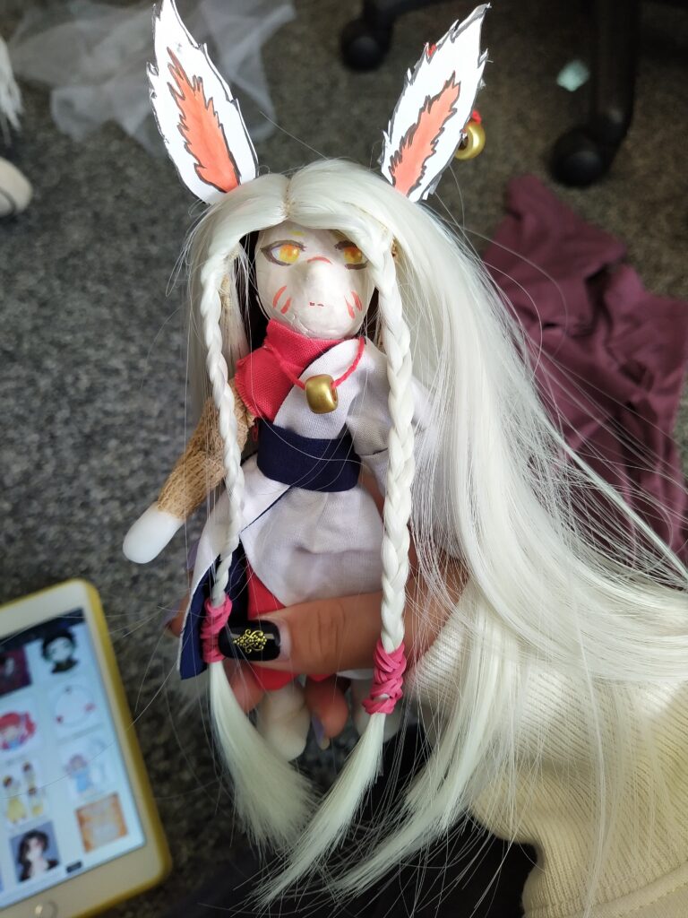 A photo of the finished puppet. She has two fox ears at the top of her head, with a bead meant to be a bell hanging from the left one. Her hair is white and goes all the way to her feet, and it's braided by the sides of her face. 