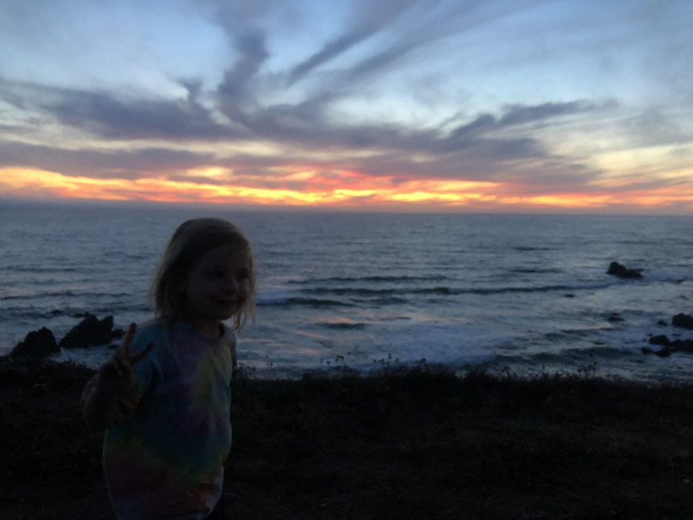 The image is of a young girl holding up a peace sign. She stands in the bottom left corner. Behind her a sunset over the ocean with yellows, reds, and blues shines. The ocean underneath the sunset has calm waves with a blue-grey color.