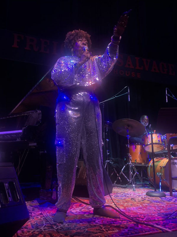 Faye Carol is on a stage in a sparkly sequined sweatsuit. She is holding the microphone up to her mouth and singing while pointing out into the crowd.