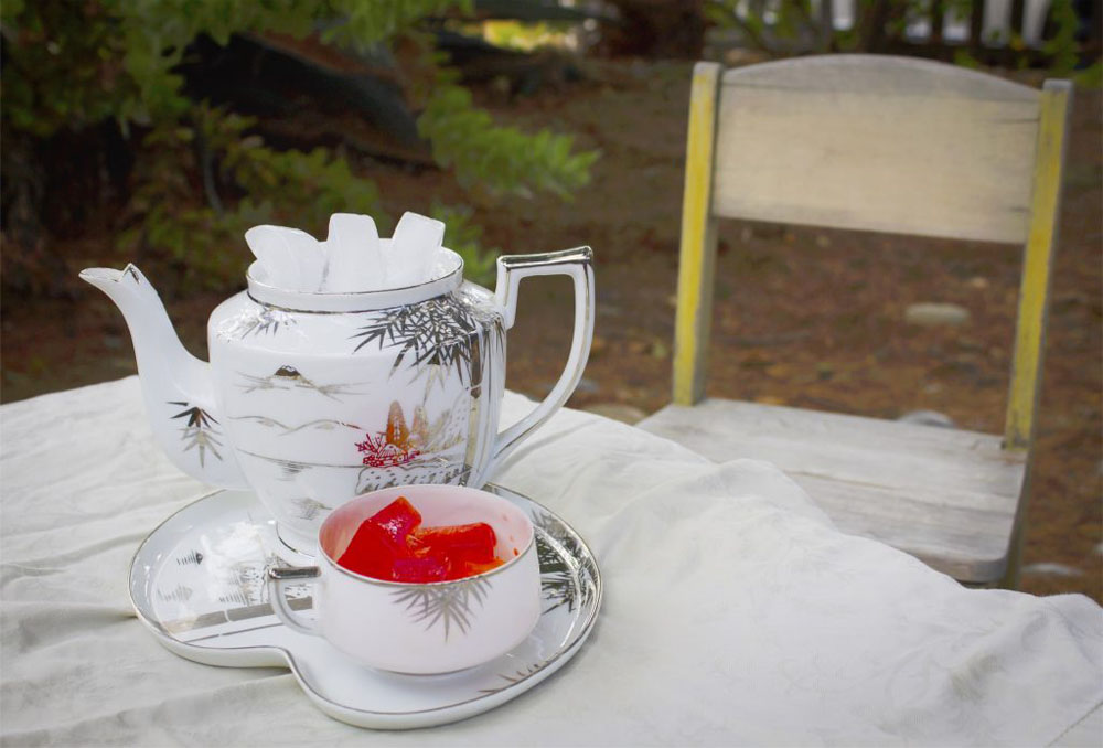 The photo is of a teapot full of normal ice next to a teacup of red and yellow ice on a tea tray. It sits on a table and a little children's chair sits empty in the background.