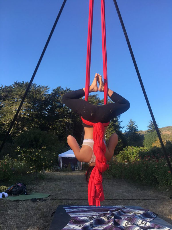 The backside of a girl upside down legs bent with feet touching. A red silk is wrapped around her suspending her in the air.