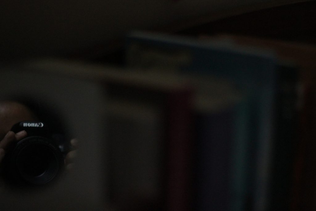 Photo of my reflection in a small mirror holding my camera, with a blurred background of different books lined up.