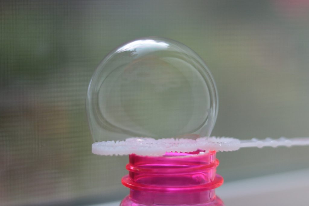 A photo of a bubble on a bubble wand with a window in the background. The color of the container with the bubble solution is hot pink. 