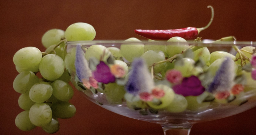 A bowl of green grapes with a red chili on top. The background is brown wood. The bowl is made of glass and has purple and pink flowers painted on it.