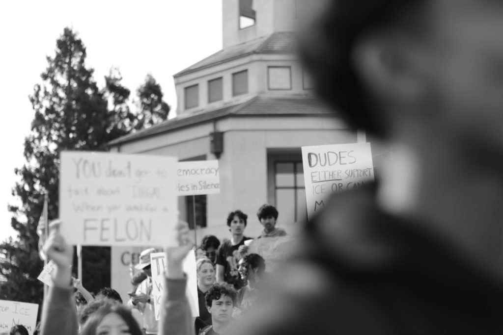 The photo is black and white. There is a figure in the right of the frame and students behind them holding signs protesting against the Trump administration and ICE. 