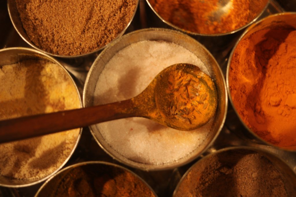 A photo of small metal tins with different spices. There is a wooden spoon in the center. 