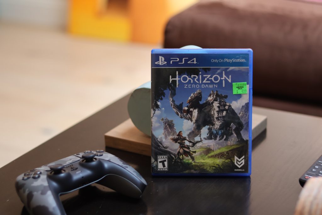 A picture of the game Horizon Zero Dawn, on a black coffee table alongside a ps5 camo controller