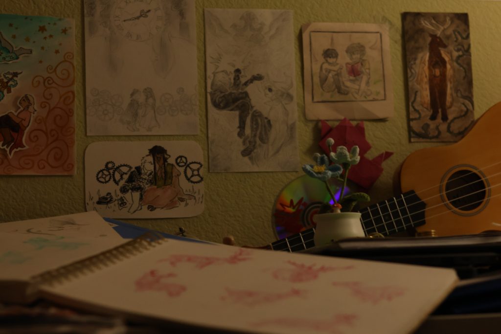 A close up photo of a person's desk and wall taken in dim, warm lighting. A ukulele sits on the desk, and on the wall are many drawings made with either pencil or watercolor. drawings in crayon are visible in the open sketchbook on the desk. 