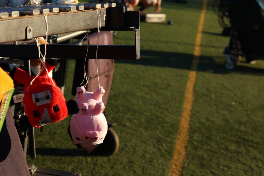 A glockenspiel, mounted on a vibraphone, takes up the left half of this photo that was taken on a football field in the afternoon. hanging upside down from the glockenspiel's keys are three stuffed animals of creatures from the game Minecraft: a orange sheep with it's Minecraft logo tag still on it, a Mooshroom cow, and a pink pig.