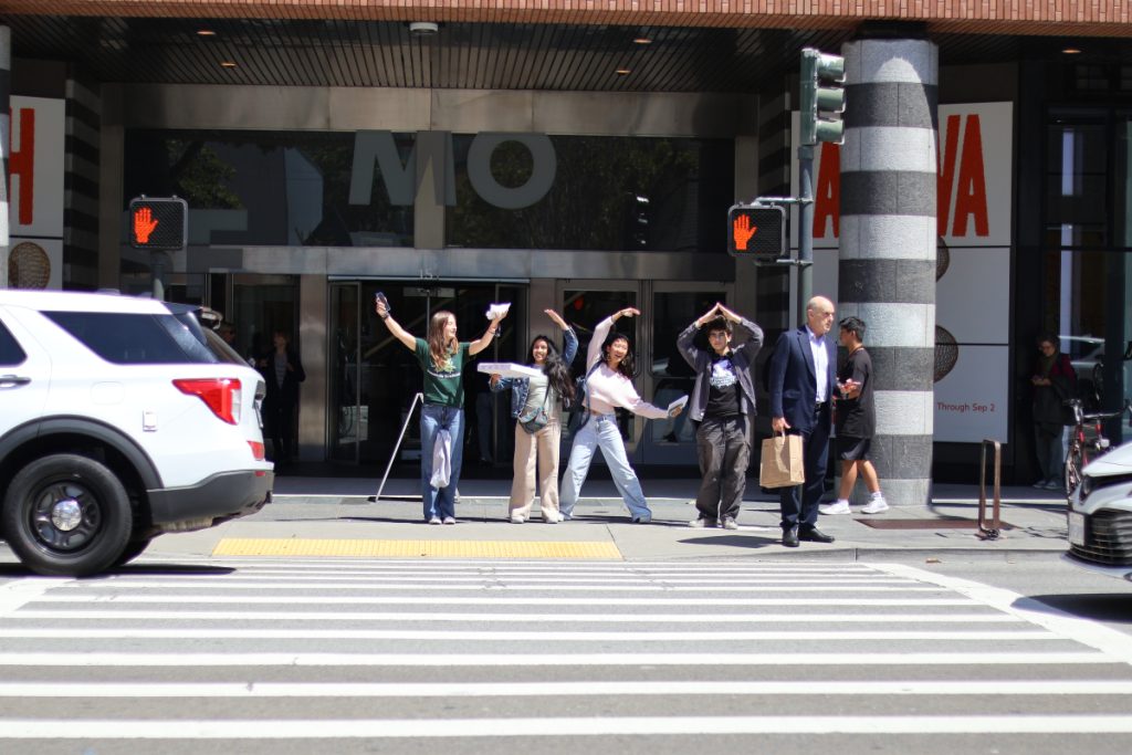 Four teenagers standing across the street from the camera use their arms to spell out the acronym YBCA.