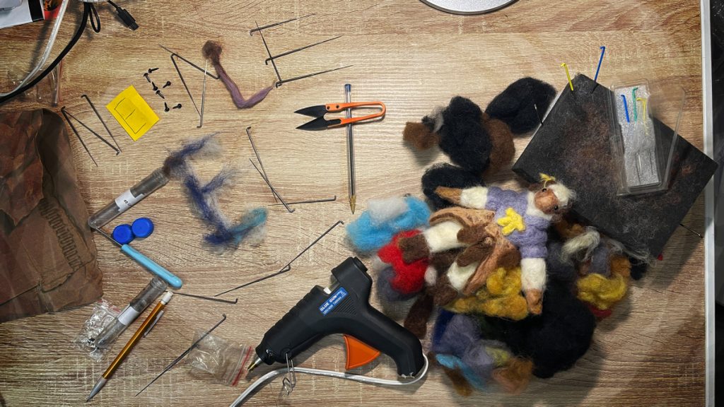 needle felting supplies and a felted figurine laid out on a desk. supplies are arranged to spell out the words "needle felt".