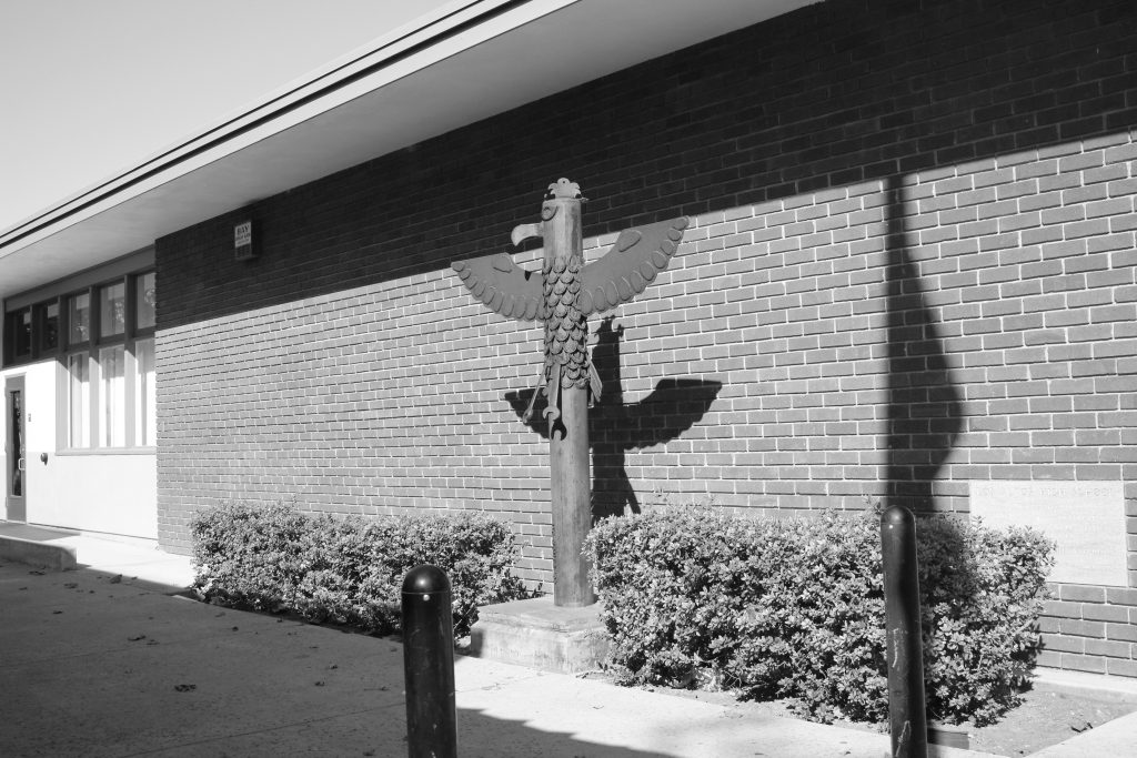 This photo shows a totem pole in the front of a high school with bushes around it. The entire shot is in black and white and there are two smaller black poles in the foreground of the shot.