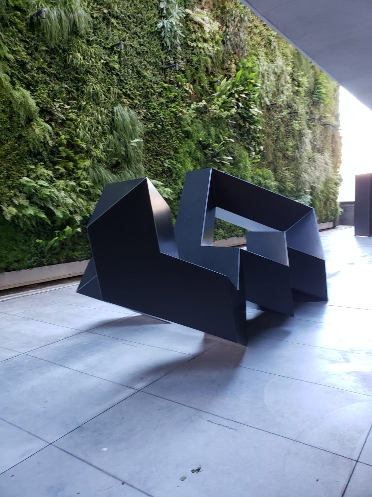 The main subject of the photo is a sculpture in the SF Museum of Modern Art. It is taking up the main part of the frame, and there is a grass wall in the background. The sculpture is black and is very rigid.