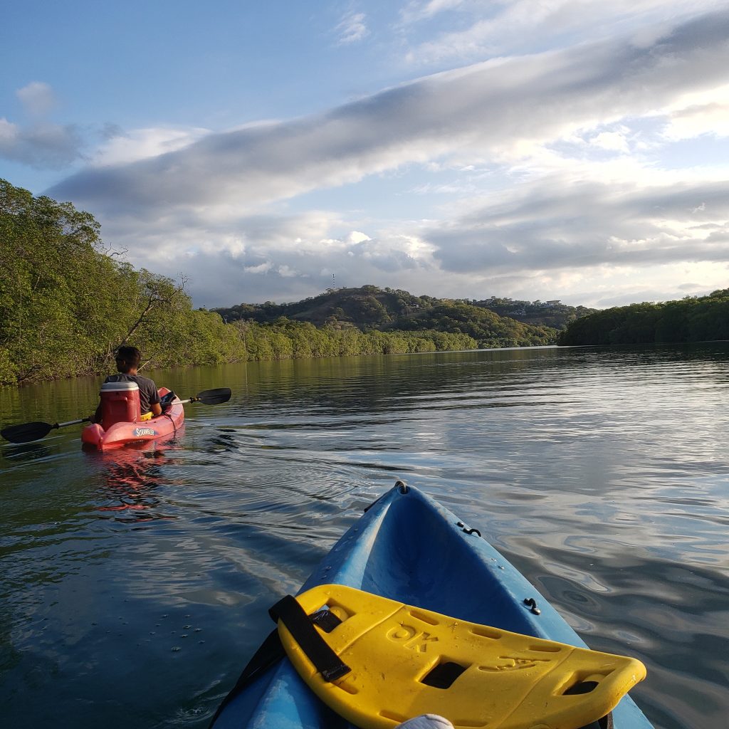 This photo shows two kayaks going down a river. A blue kayak is in the foreground and a red kayak is in the background, along with a hill and trees surrounding the river.