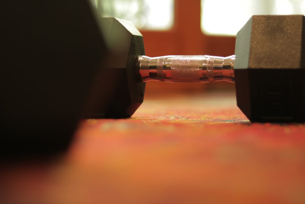 This picture shows a dumbbell in the background, with a blurred out dumbbell in the foreground. They are both resting on a red carpet.