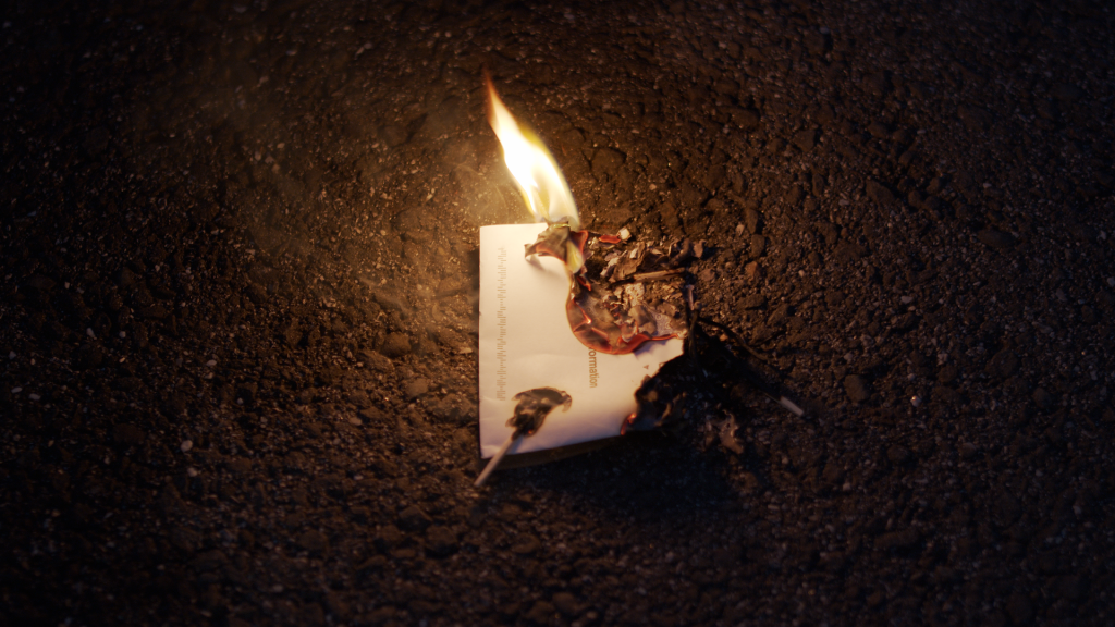 This photo shows a letter being lit on fire. There are matches on some parts of the envelope, and the background of the photo is blacktop.