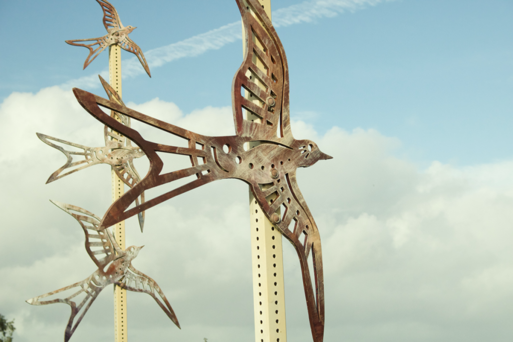 A large metal bird in the center, with 3 smaller metal birds behind it, with a blue sky surrounding them.