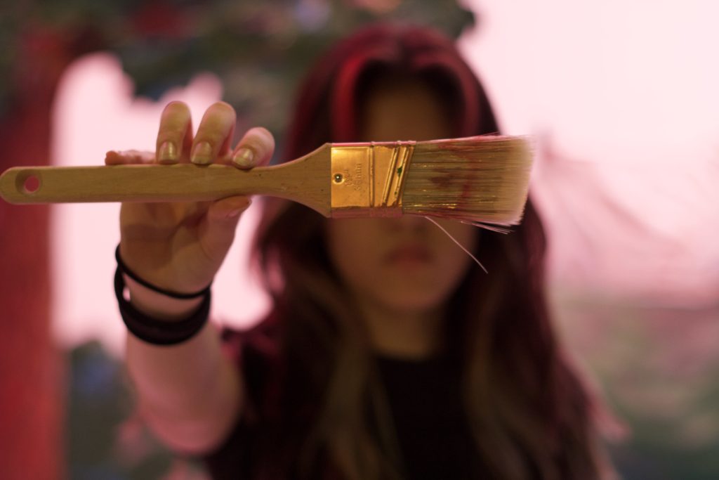 Athena Driesen standing in front of the set of the school play (that she designed), holding out a paintbrush towards the canvas to cover her eyes.
