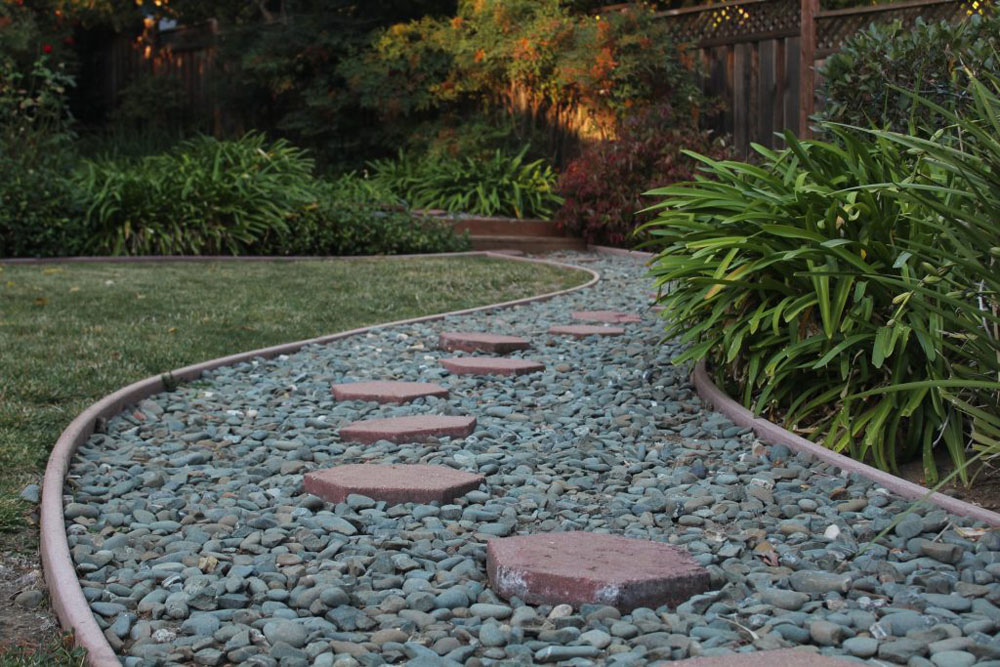 This photo is outside. The focus of it, the pathway filled with gray pebbles and discolored red stepping stones, takes up the majority of the photo. On it sides are grass and plants as well as a fence. 
