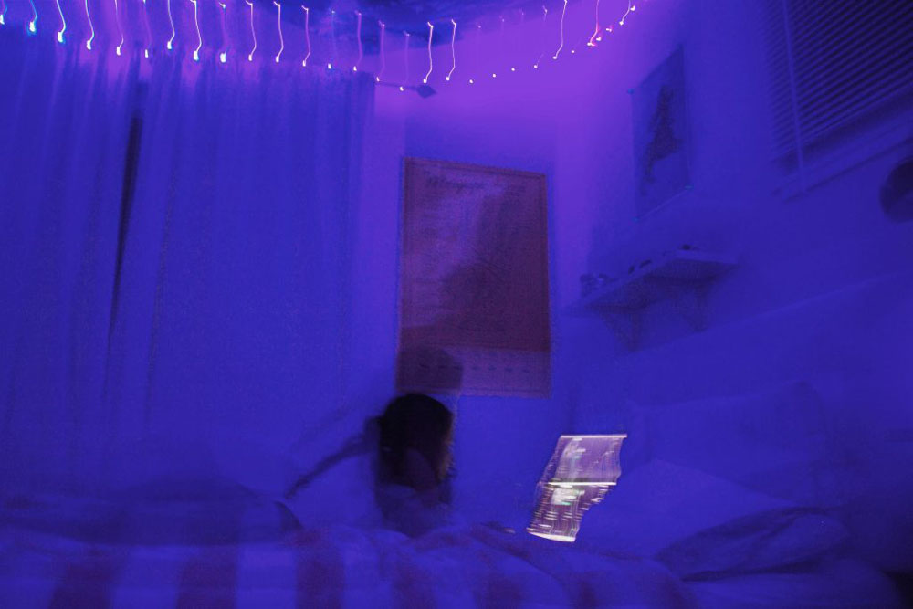 Picture of a girl sitting on a bed looking at a computer. The room is dark and is blue/purple. The picture is in motion.