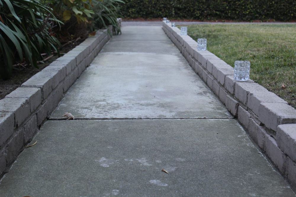 Picture of a gray pathway with light gray bricks on either side of it. The left side has bushes, the ride side has grass.