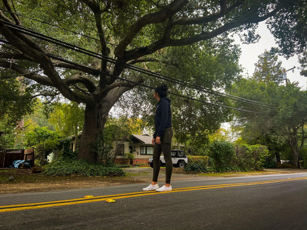 A girl wearing a blue sweatshirt and green pants standing in the middle of the road looking up at a big tree that takes up the majority of the background. A small white house can be spotted in the distance and other trees surround the area. 