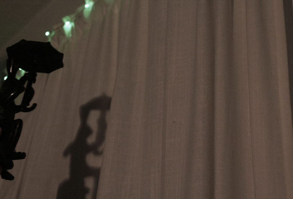 A picture of a shadow of an object that is partly in the frame that is shaped like a frog holding an umbrella- reflected onto a white curtain with green LED lights at the top. 