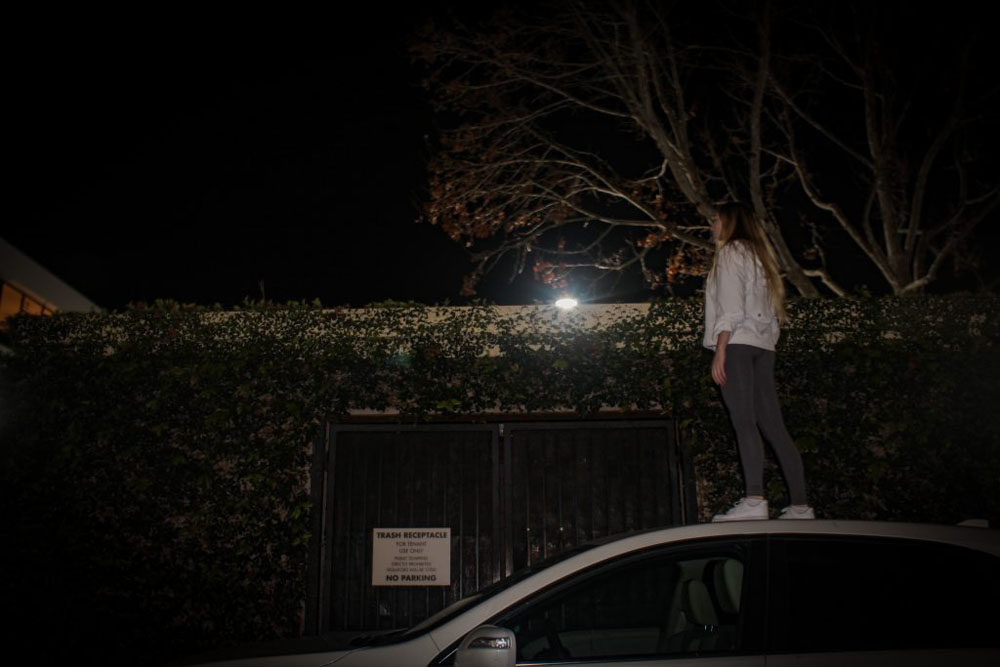 A girl in a white jacket and grey pants standing on top of a white car in front of a white dumpster with leaves all over it and a tree in the back in the nighttime.
