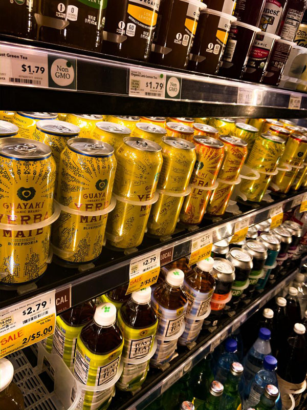 Picture of rows of drinks in a grocery store fridge. The main focus is a row of Yerba Mate drinks that are primarily yellow.