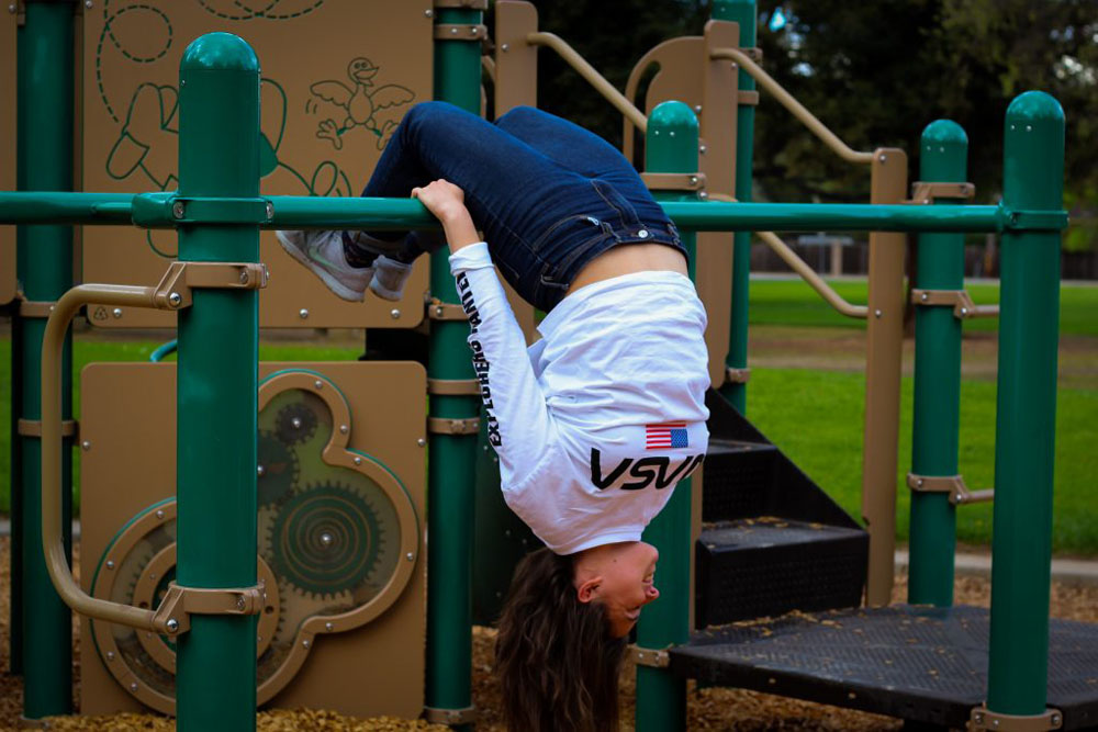 Girl in a white shirt and blue jeans hanging upside down from the green monkey bars on a playground.