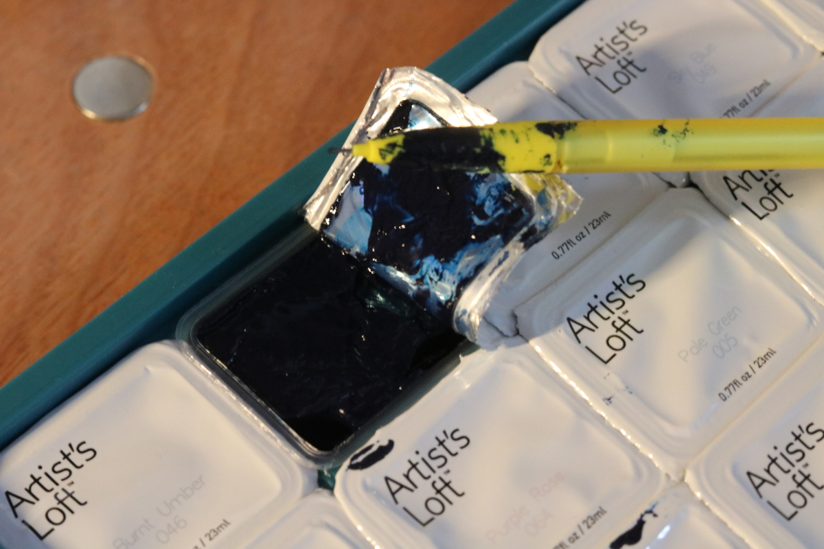 A picture of my jelly gouache palette. The Prussian blue capsule is open and a yellow mechanical pencil is soaked in it whilst keeping it open. 